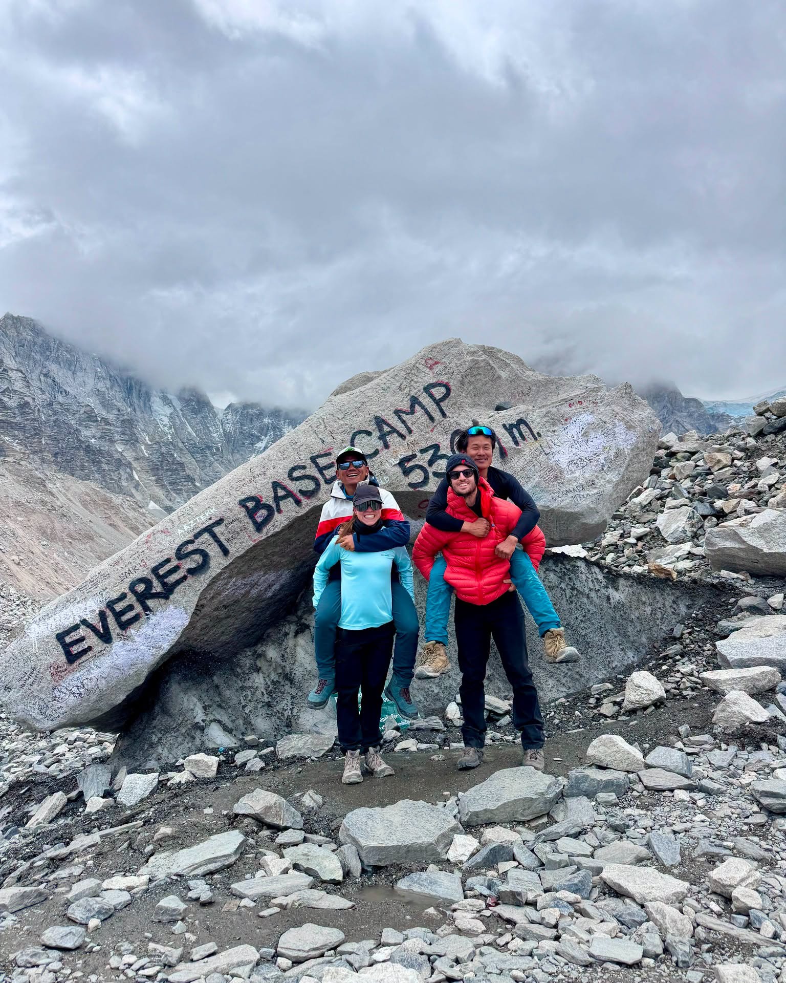 Four trekkers celebrating at Everest Base Camp 5364m marker stone with Himalayan mountains in the background, Nepal
