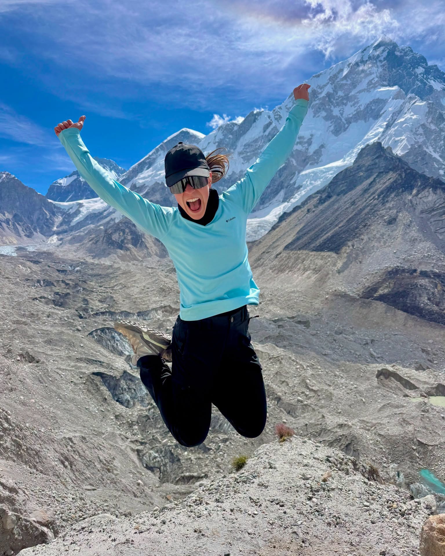 Trekker jumping with arms raised in celebration against a backdrop of snow-capped Himalayan peaks including Nuptse on the Everest Base Camp trek